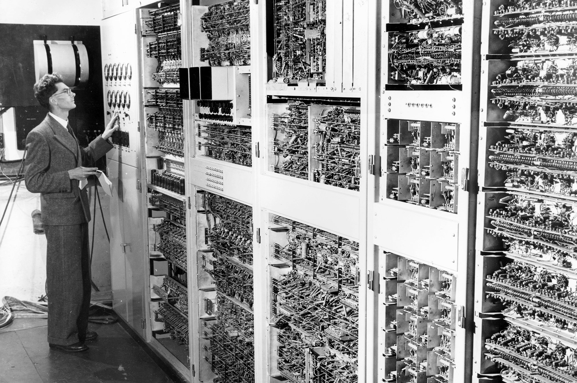 Black and white photo of a man in a suit operating a very old computer which is several cabinets in a row with many wires, switches and dials. 