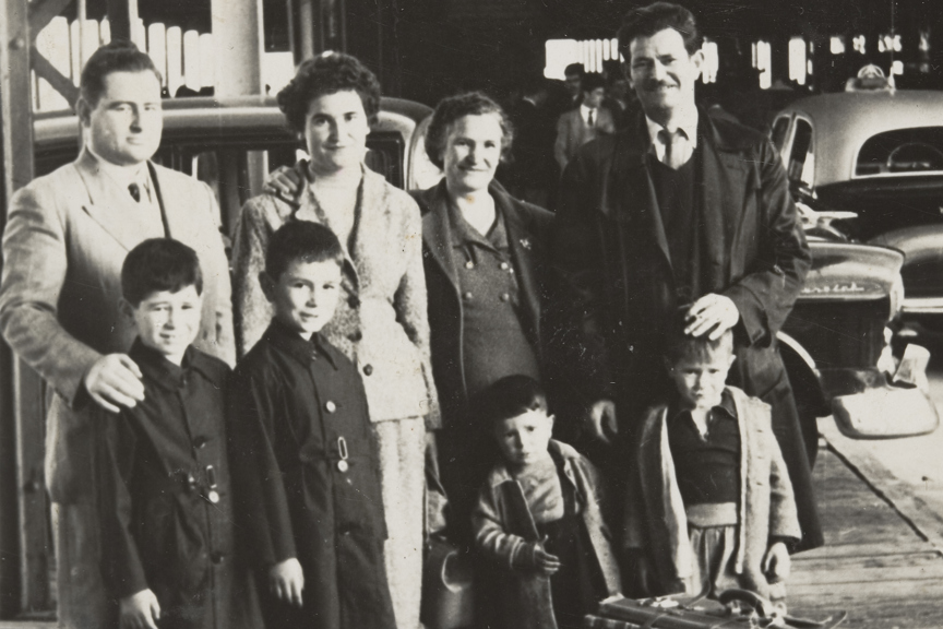 Extended family standing around suitcase on Station Pier, under a large roofed structure. Two men, two women, two boys and two infant children. There is a large suitcase in the foreground with rugs strapped to the side. Cars are parked in the background.