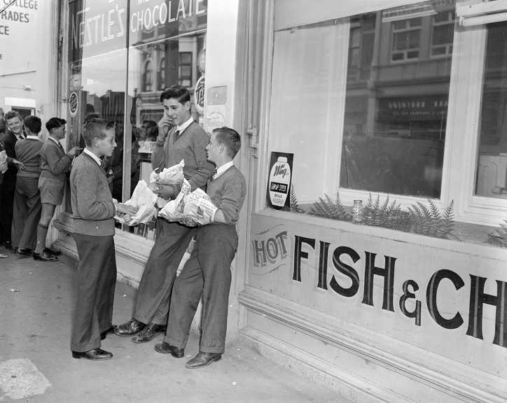 School children outside a takeaway store eating food wrapped in newspaper. 