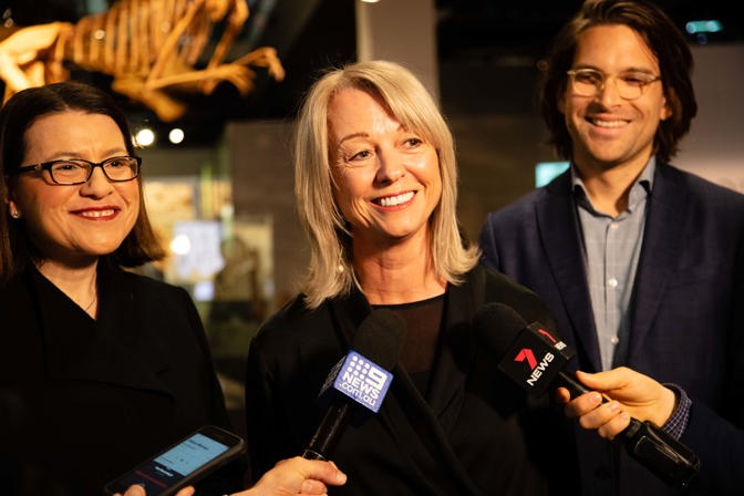 A woman speaking in to news microphones. A woman stands to her left and a man to her right