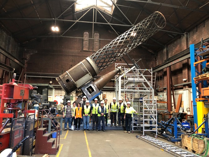 Group of people wearing hi-vis vest stand in front of a large metal cylinder in an engineering workshop 