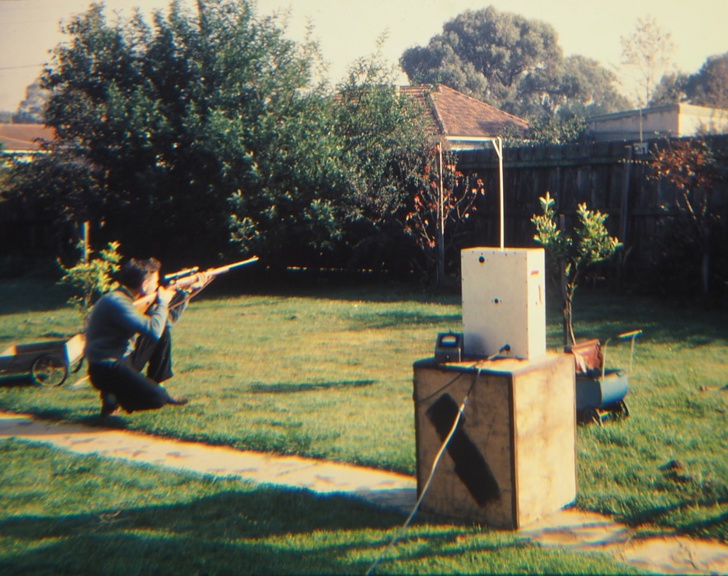 A young man in a suburban backyard with a rifle. 