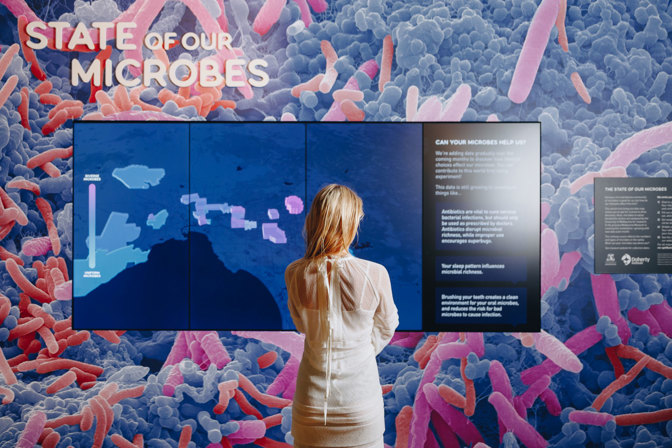 Woman standing with her back to the camera wearing a cream dress view a large exhibition panel.