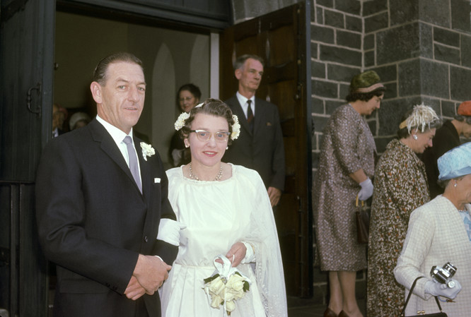 Bride and groom outside church to the left of image and facing camera. Some guests in background outside and inside the church.