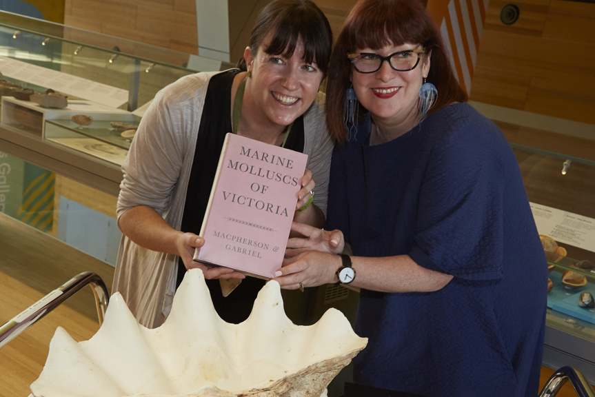 Two women hold a pink book in front of a large shell. 