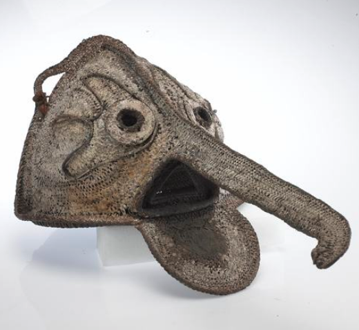 A fibre figure resembling a head coated with natural pigment. It has gaps for the eyes and mouth and an elongated nose.