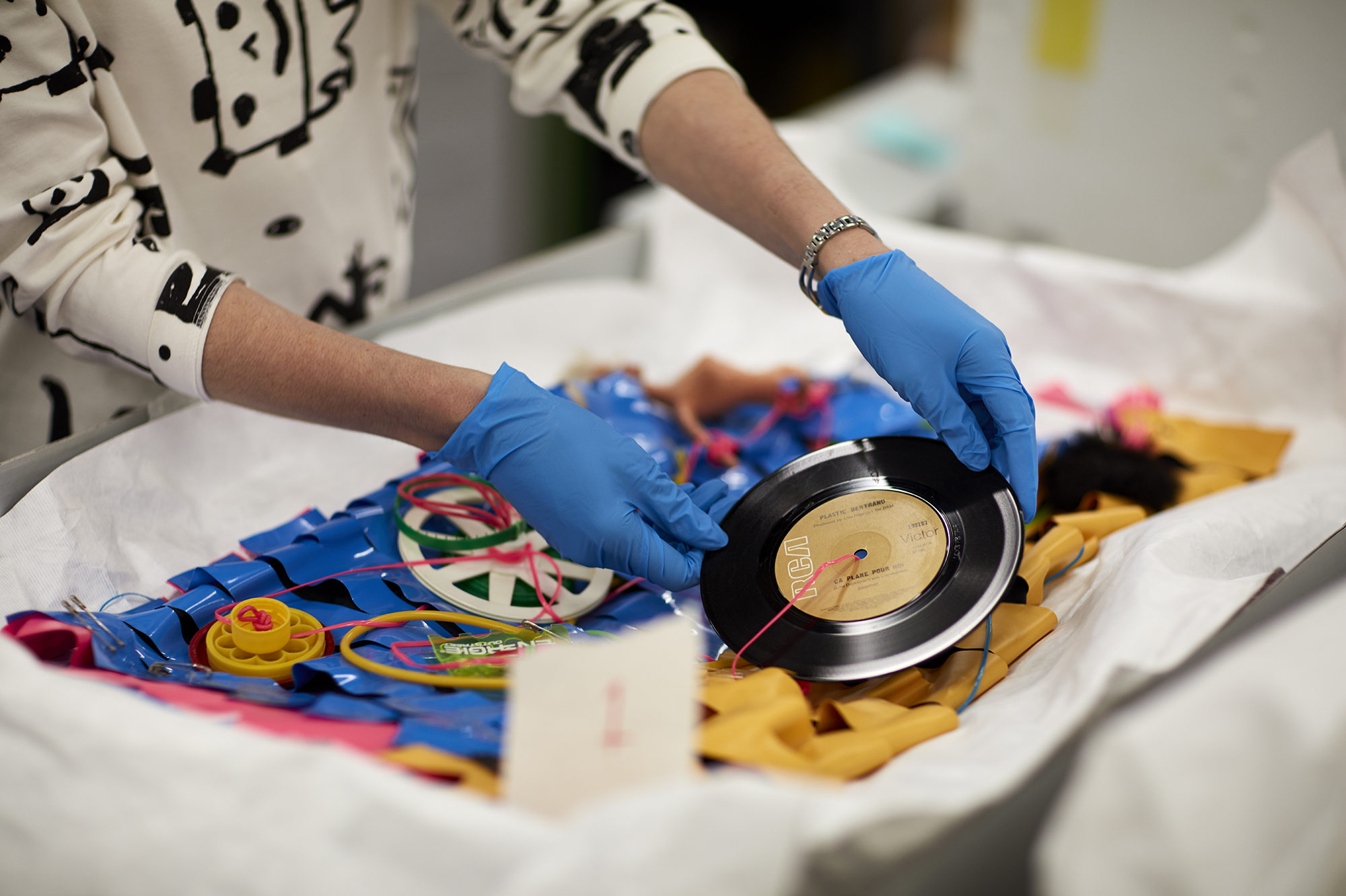 Conservator, wearing blue gloves, handling Jenny Bannister's blue plastic 'Je Suis Mod' dress, holding a vinyl record decoration.