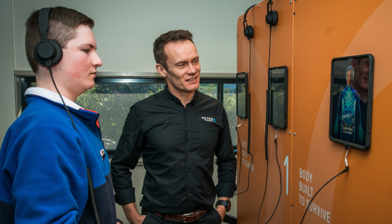 A Road to Zero presenter stands beside a student who has headphones on and is watching a screen on a wall