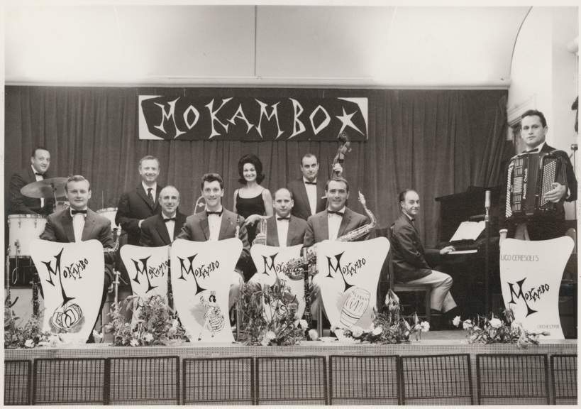 Eleven Mokambo Orchestra band members, including a female singer. Band leader Ugo Ceresoli with accordion at far right. There are six music stands standing in front of band members across the front of stage.