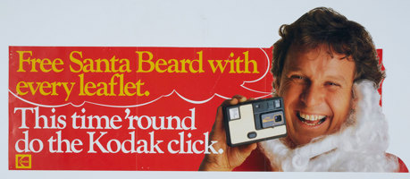 White, red and yellow poster with an image of a man with a santa beard pull below his chin holding a small camera. Text " Free Santa beard with every leaflet. This time "round do the Kodak click"