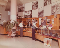 People standing in a photographic shop. Two male shop assistants stand behind the counter while two customers stand with them in front of the counter.