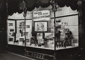 A shop front display case. Cameras and accessories on floor of case, plus posters and a large sign promoting Kodachrome colour film.