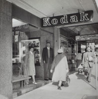 People walking along a footpath and standing in the doorway of a photographic shop.