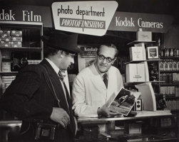 Man standing behind a counter in a photographic shop showing a photograph of a woman to another man standing on the other side of the shop counter.