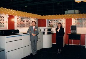 Woman and man with yellow nametags standing in a trade show/ conference stand with business imaging equipment around them.