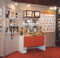 Tiered carpet board panels with photographs and display panels mounted on them. Red and white table with Perspex display cases above filled with products and labels. Cubed light boxes at top of display.