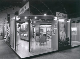 Exhibition display stand viewed from its corner showing lighting and steel roof supports above the stand, and various divided display areas.