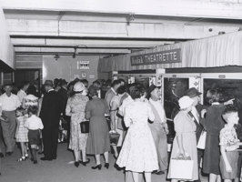 Crowd of women, men and children outside 'Kodak Theatrette' with photographs mounted in light box on right wall.