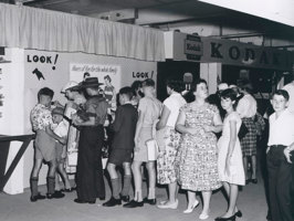 Two rows of children, with the occasional adult, lining up to look into wall mounted display boxes. 