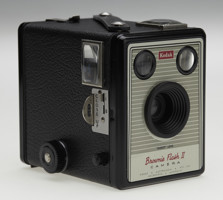 Small box camera with fixed lens and two view finders. Camera body is black with its front face featuring grey stripes on silver with red and black text.