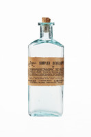 Glass bottle with cork stopper and paper label on the front with printed text in black ink. Bottle is empty of contents but there are some powdered residues on the cork.