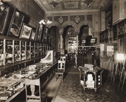 A shop interior, display counters and cases. Photographs hanging on walls. Chairs for clients to be served in. There are columns at the rear of the store with elaborate paint and plasterwork above them. The letter 'K' is painted in the centre of wall and ceiling friezes.