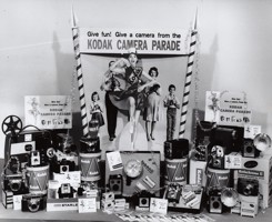 A display featuring a 3D cardboard poster of a family of four behind a marching girl. Camera products and accessories displayed in window. Some items placed on drums.