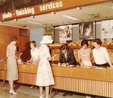 Five women and one man in a shop that sells photographic materials. Three of the women and the man are behind the shop counter. Two women are standing in front of the counter.