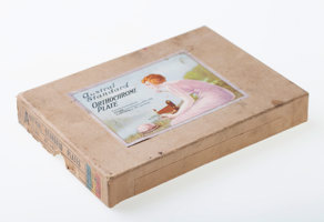 Brown cardboard box lid with label adhered to top face. Label features an illustration of a woman in a pink dress with pin-curled, short hair, holding an extended folding camera outdoors.