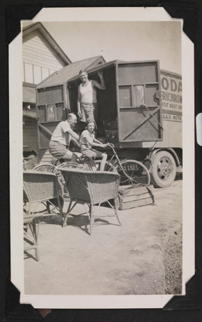 Man and a child riding a bicycle with a man standing in the back of an open delivery van behind them.