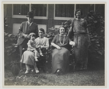 Family posing for group portrait in garden outside house. Shrub behind them. Two women are seated, one with a child on her lap. Man in hat stands at left and younger woman in dress stands at right.