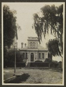 Exterior of large house taken from across the street showing the road, street lamps, drains, trees, and neighbouring properties. The house has a slate roof, at least 3 chimneys, a large entry stairway with balustrades and a tower with balcony above the front door. The front fence is wrought iron.