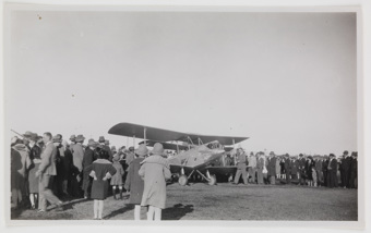 Large crowd gathered behind a small biplane parked on a grassy lawn
