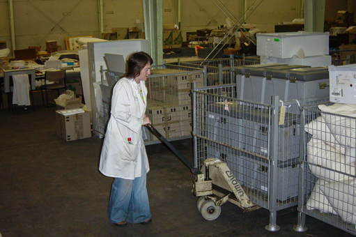 Woman in jeans and white laboratory coat pushing pallet of plastic crates in a warehouse