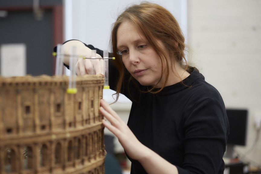 An auburn-haired woman works on a cork model of a Roman Colosseum