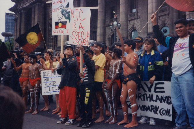Group of people standing on the steps of parliament house holding placards and aboriginal flags