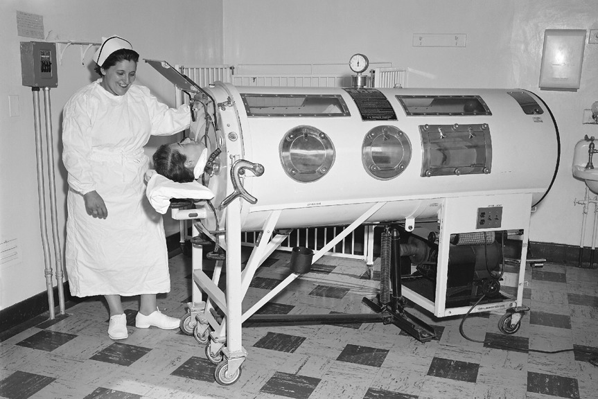 A man encased in a large metal box. His head is exposed and a nurse is smiling down at him