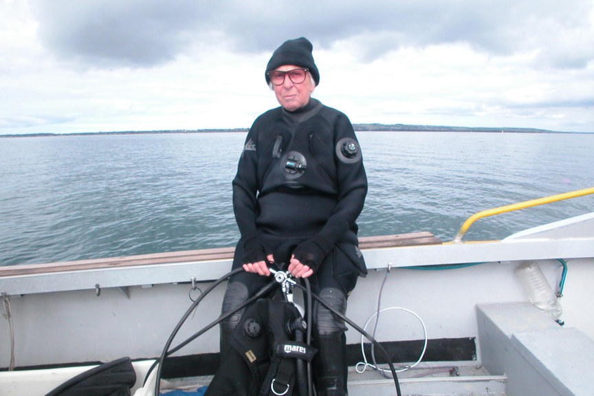 an elderly woman wearing a wetsuit and sunglasses sitting on the edge of a boat on the water