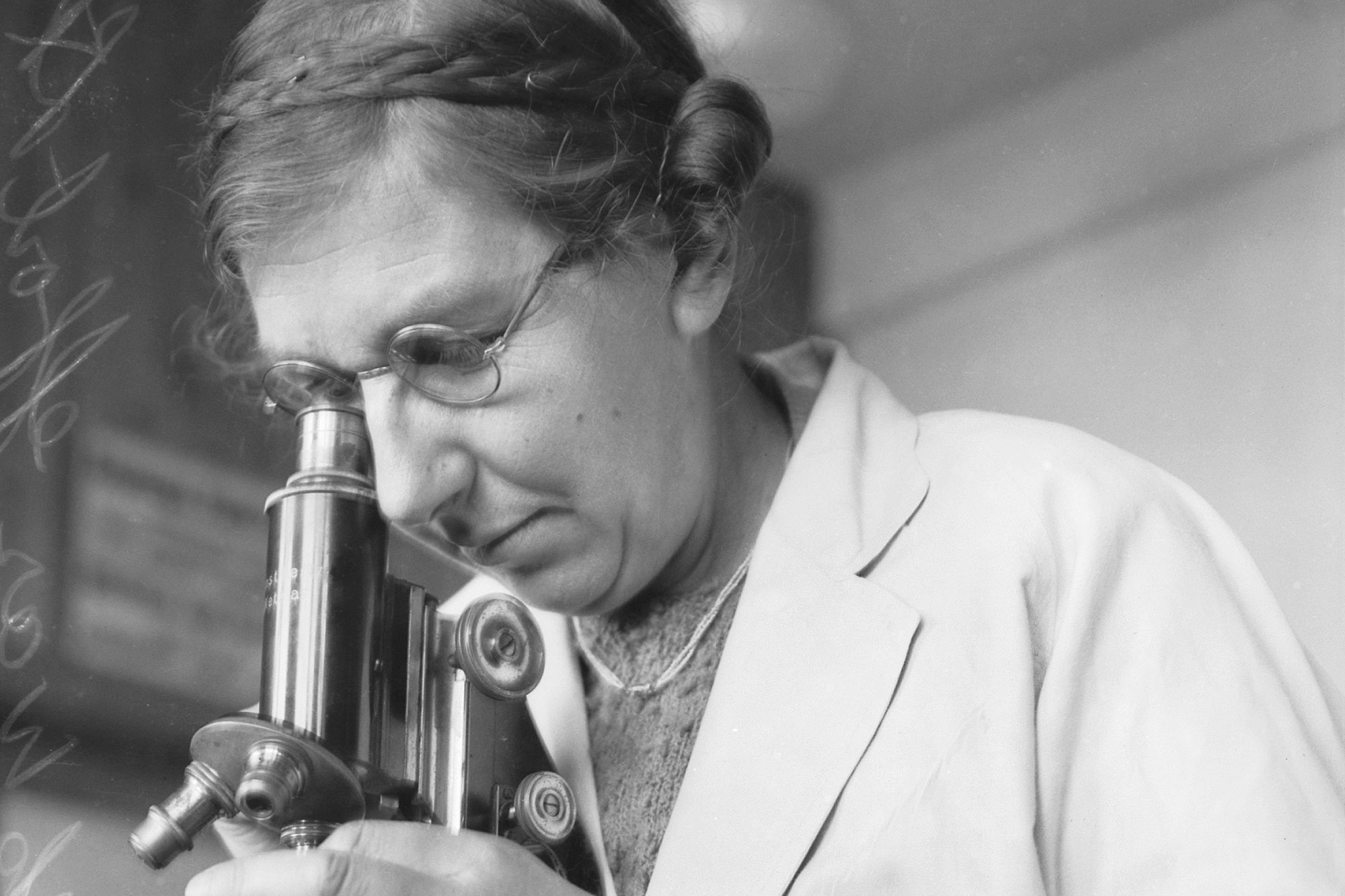 a black and white photo of a woman wearing glasses and a white coat, looking into a microscope