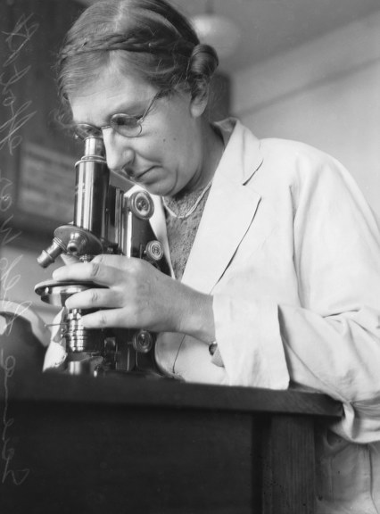 a black and white photo of a woman wearing glasses and a white coat, looking into a microscope