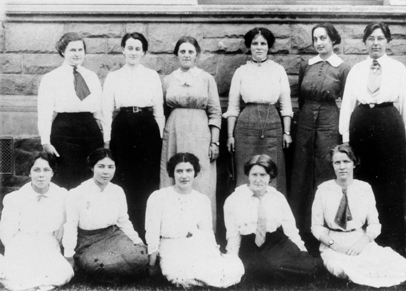 a black and white photo of 11 young women pose in front of a brick wall for a photograph 