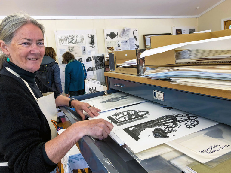 a photograph of a silver haired woman wearing an apron in front of a large drawer filled with black and white artistic prints