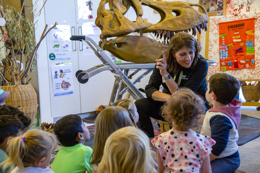 A woman sitting in front of a group of young children points at a cast of a dinosaur skull with big pointed teeth