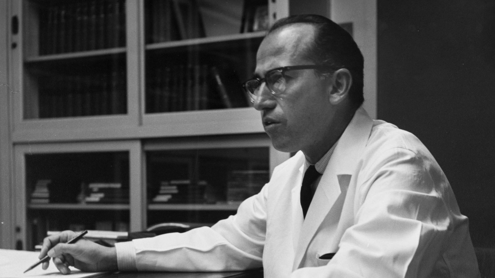 a black and white photo of a man wearing glasses and a white coat sits at a desk holding a pen 