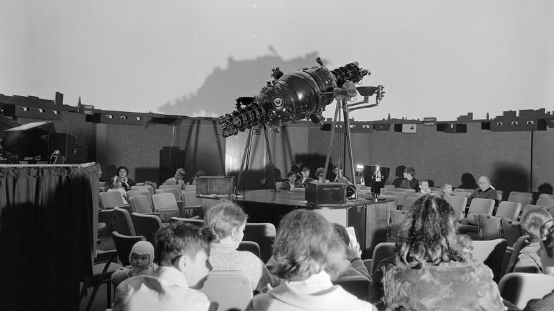 Black and white photo of audience seated in a planetarium around a large star projector before a night sky presentation