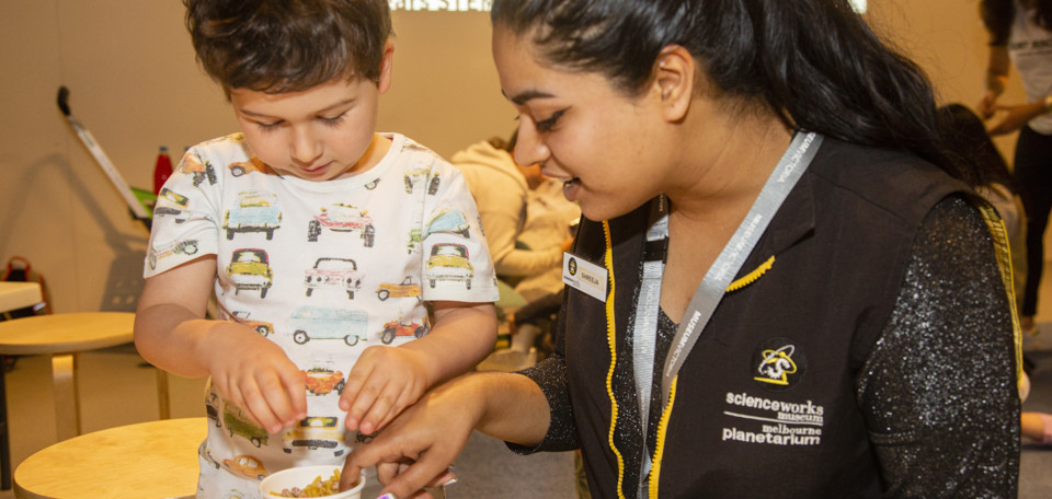 A woman with black hair in a ponytail sitting at a work table with a young boy with brown hair looking into a white cup full of things to create sound