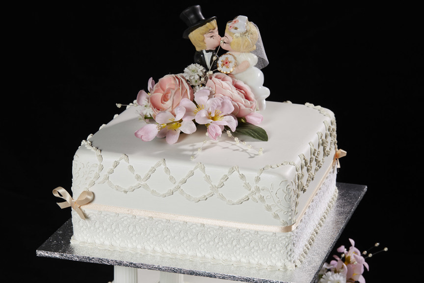 A white cake with flowers and small figurines of a suited man and woman wearing a white dress