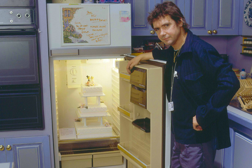 A man holds the door of a fridge open. A three tiered wedding cake sits inside.