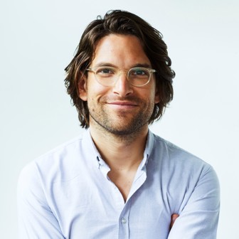 A smiling man with shoulder length dark wavy hair parted to the right, light brown round glasses and light beard. He's wearing a light blue button down shirt.