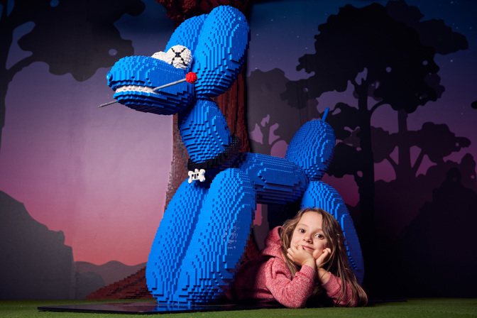 A blue Lego dog with a girl lying beneath it, head in hands. There is a woodlands backdrop.